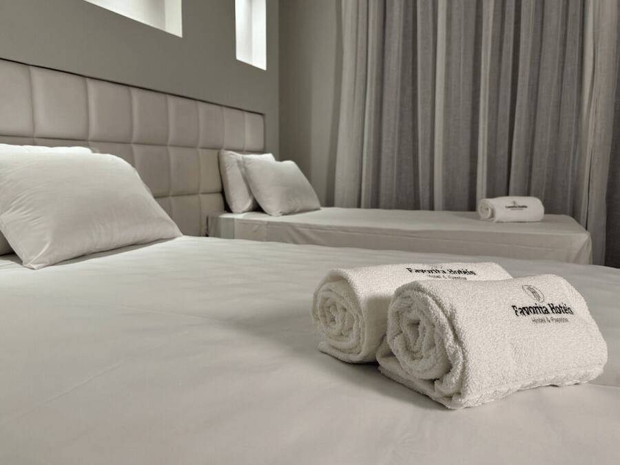 Quarto de hotel com uma cama grande, duas almofadas, uma toalha enrolada e uma mesa de cabeceira com abajur.