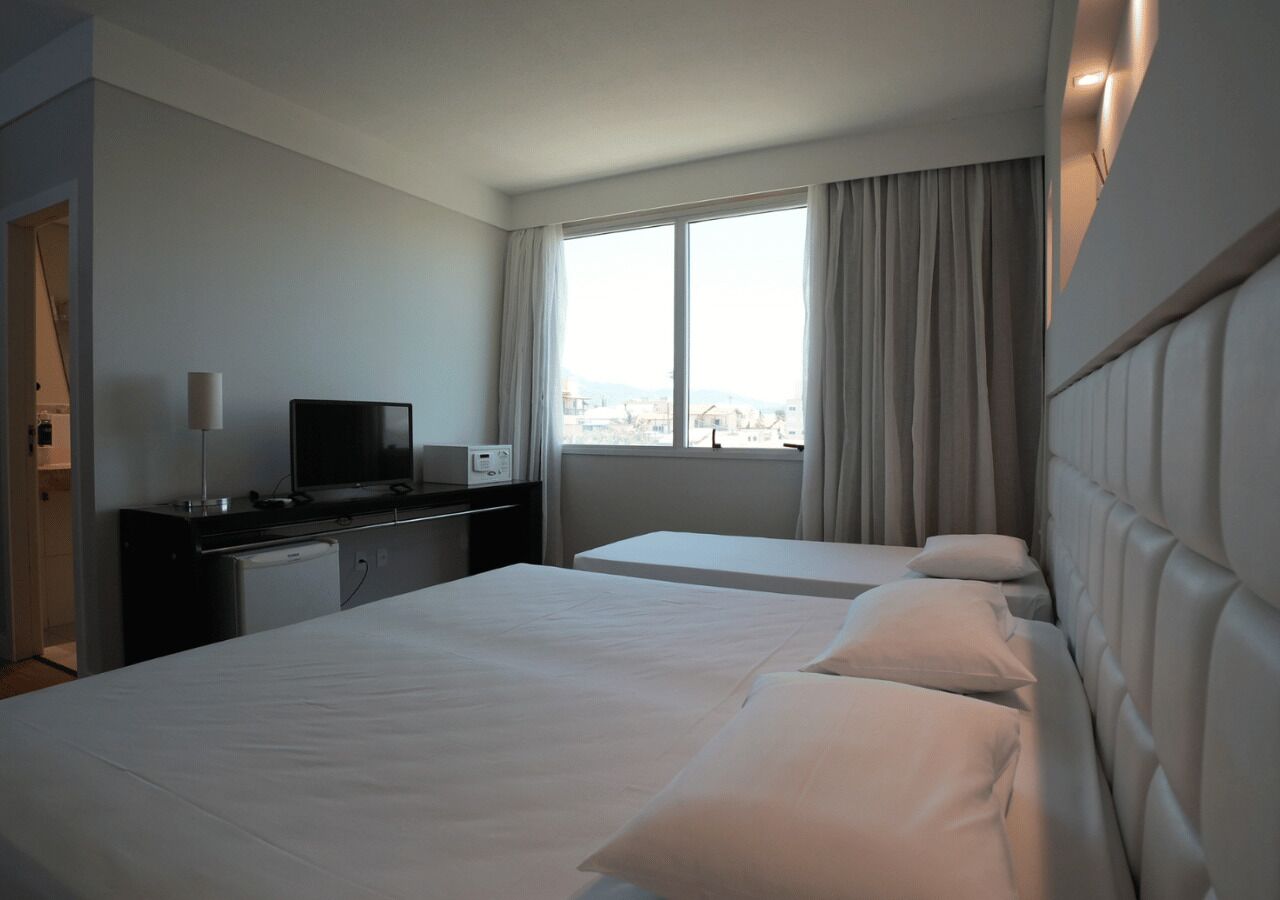 A hotel room with two beds, a desk with a television, a window with curtains, and a view of the city.