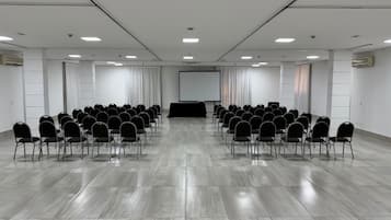 Un salón de conferencias con filas de sillas, una pantalla de proyector y un amplio ventanal.