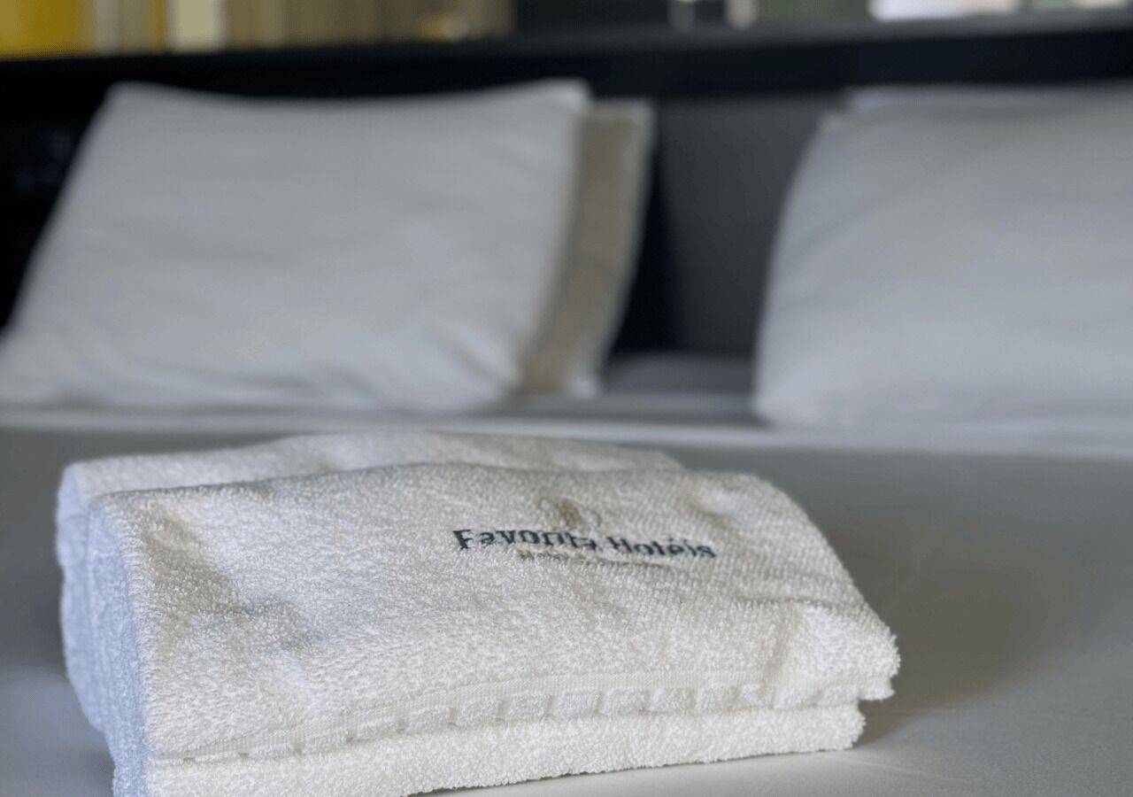 Un asciugamano bianco piegato con la scritta 'Favona Hotels' su un letto con cuscini bianchi e un poggiatesta grigio.