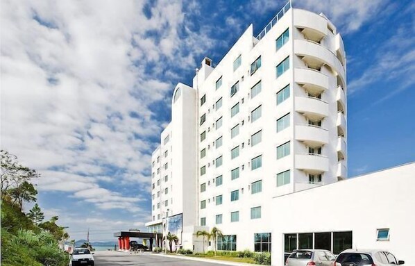A multi-story hotel with a white facade, balconies on each floor, and a parking area in front.
