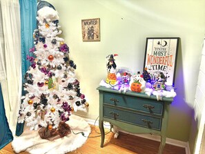 A decorated Christmas tree with ornaments and a small figurine, a dresser with a sign, and a wall with a framed picture.