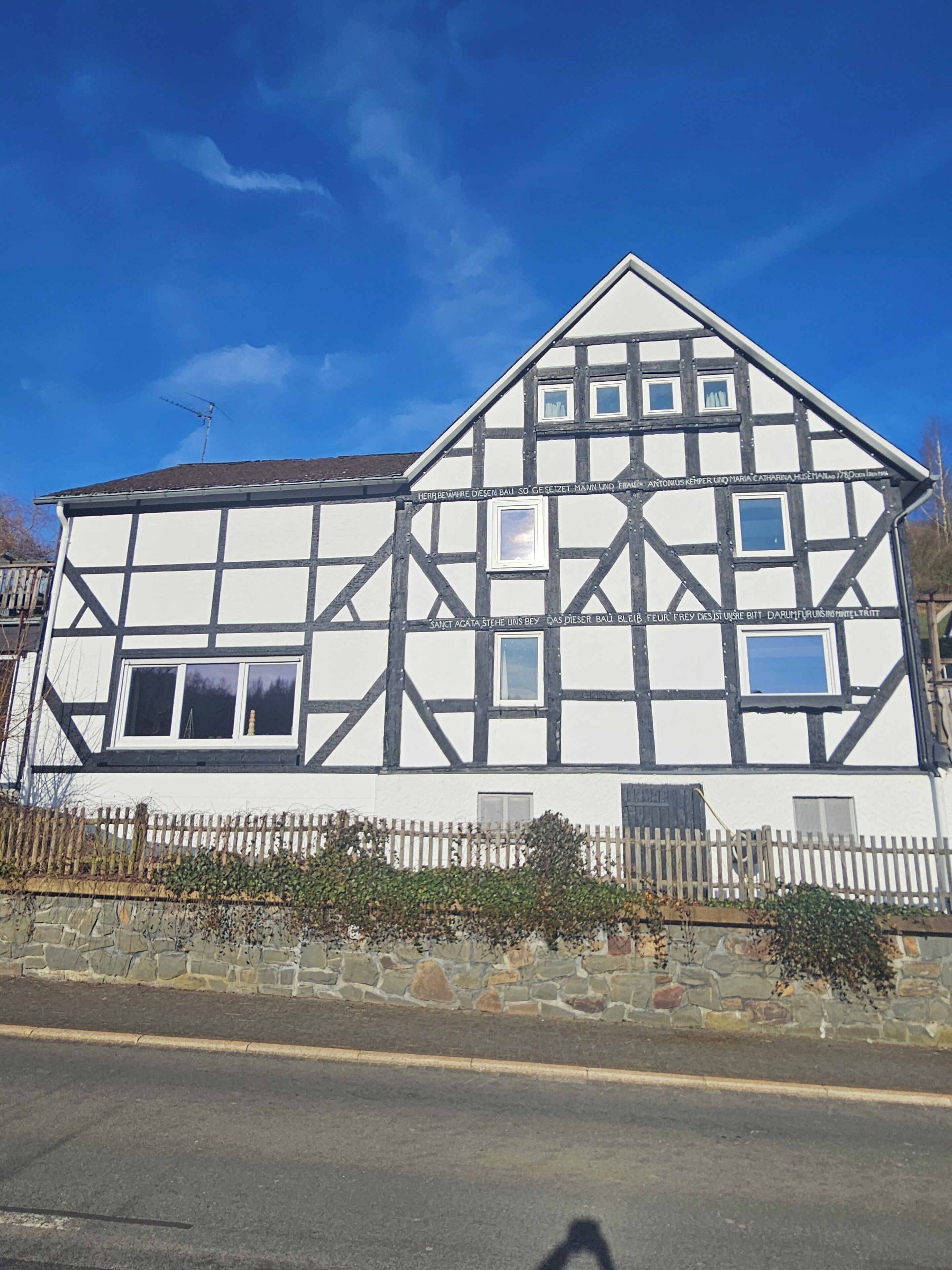 A traditional half-timbered house with a stone wall foundation, a wooden fence, and a clear blue sky in the background.