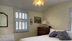 Ein Schlafzimmer mit Bett, Schminktisch, Nachttisch, Lampe, Spiegel und Fenster mit Jalousien.