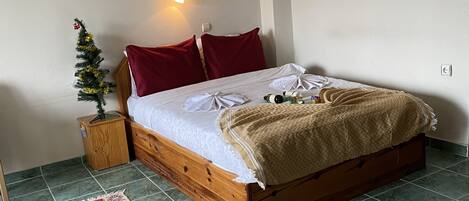 A neatly made bed with a red and white blanket, a small wooden nightstand with a plant, a red pillow, and a framed picture on the wall.