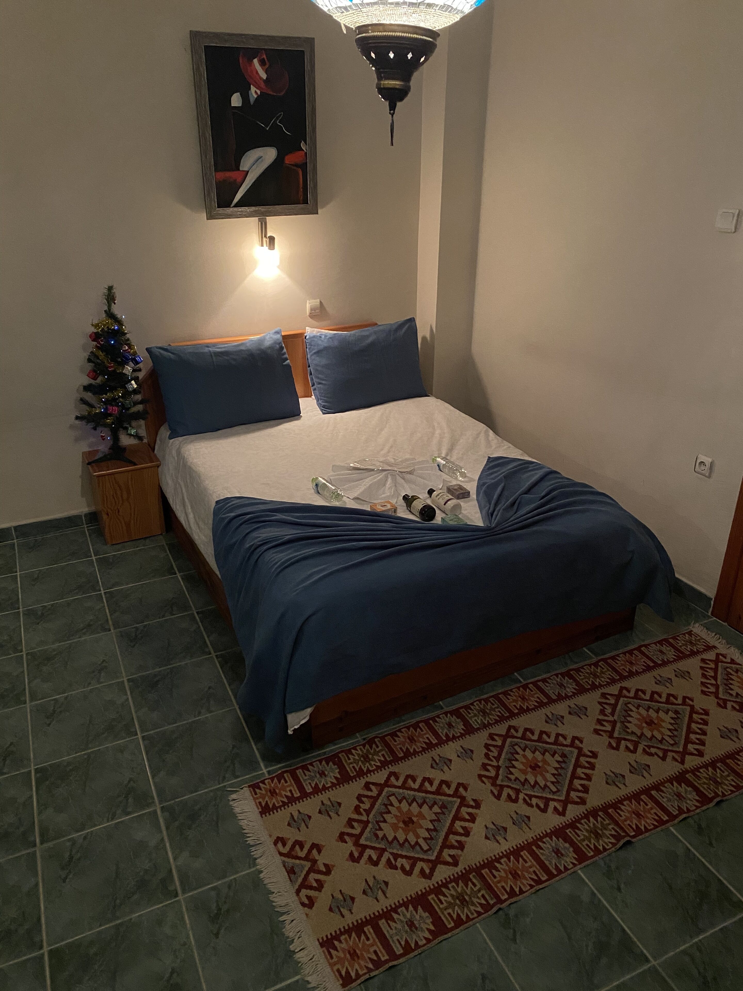 A bedroom with a bed, a small Christmas tree, a painting, and a patterned rug.