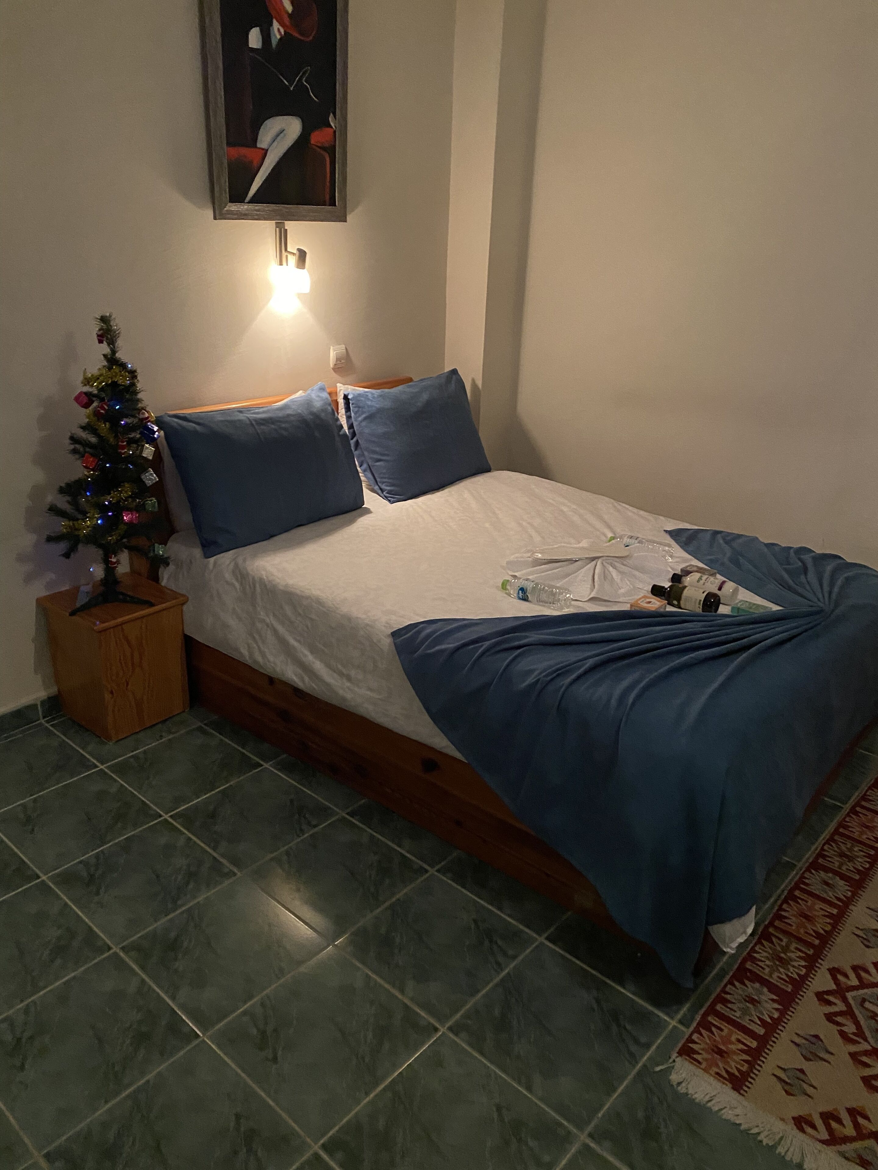 A bedroom with a bed, a small wooden nightstand, a Christmas tree, and a painting on the wall.