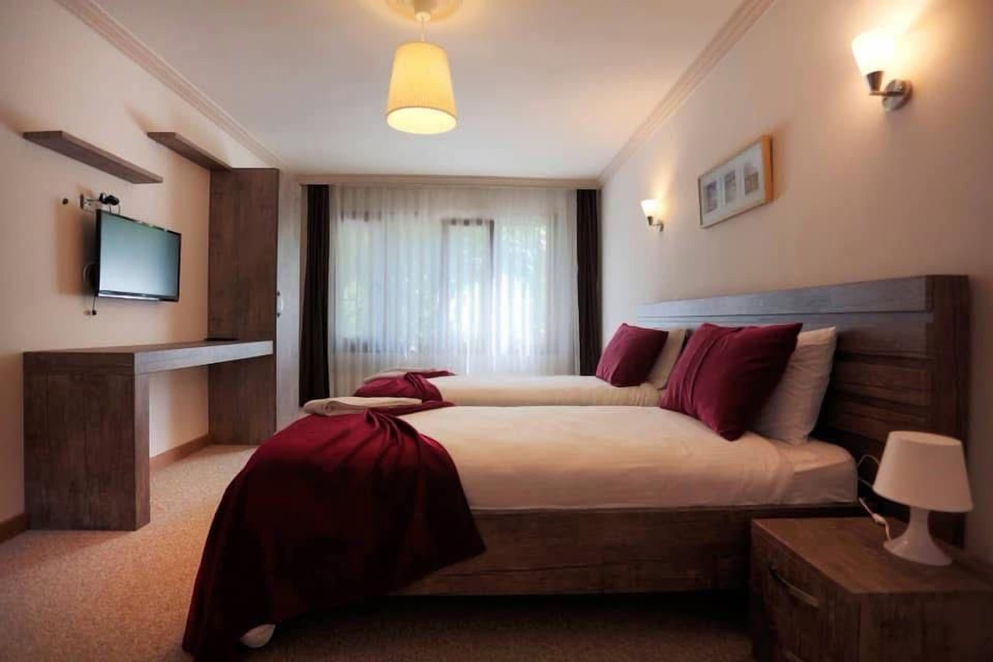 A hotel room with two beds, a television on the wall, a bedside lamp, and a window with curtains.