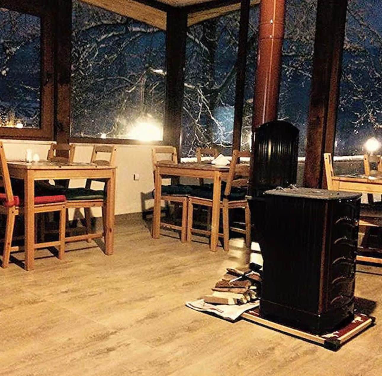 A room with a dining table and chairs, a fireplace, and a view of trees through large windows.