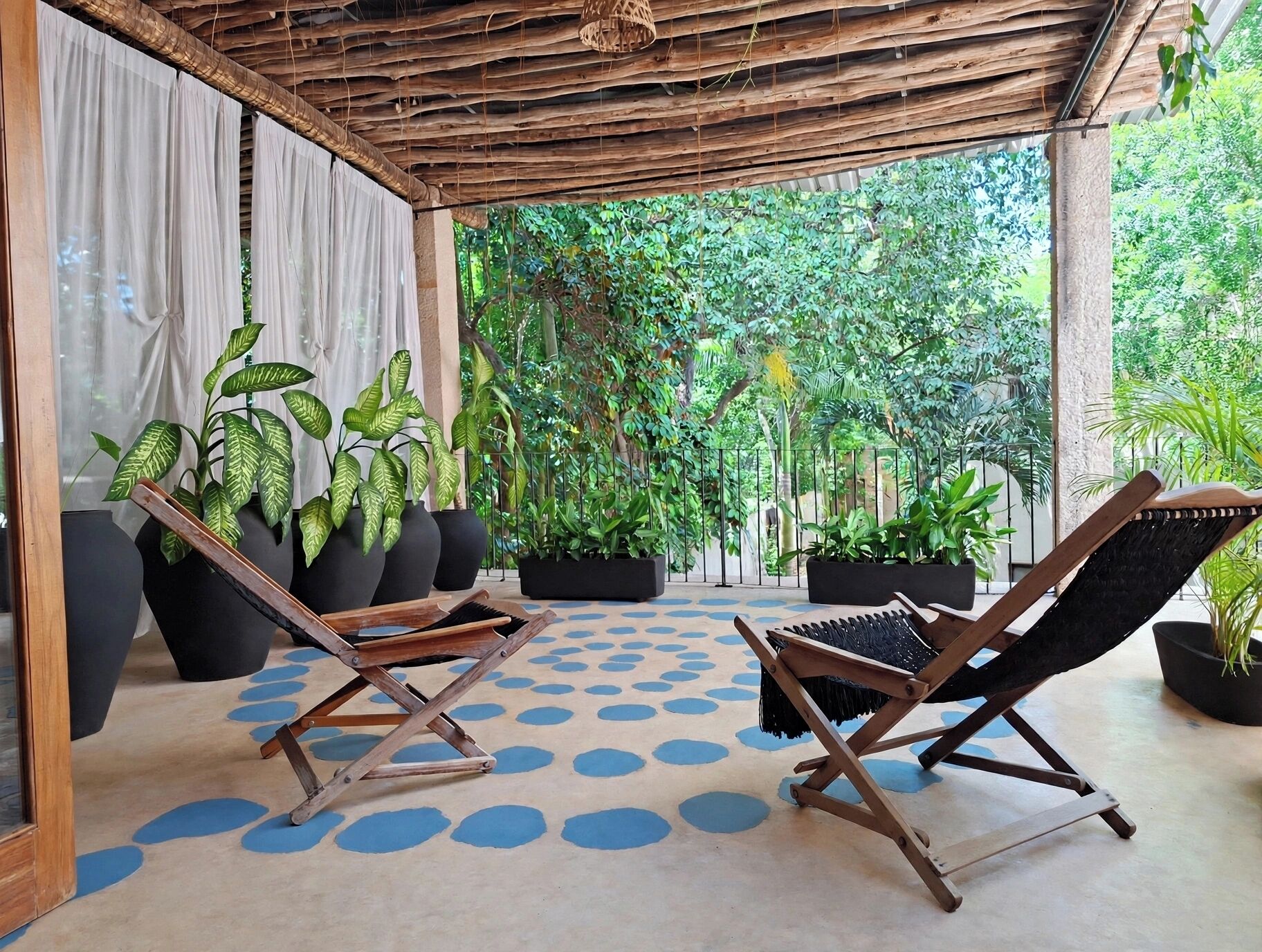 Un espacio de descanso al aire libre con techo, dos sillones de madera, piso con diseño y abundante vegetación de fondo.