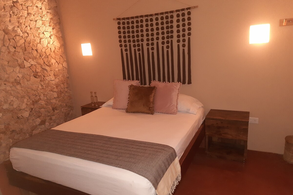 A bedroom with a stone wall, a bed with white bedding and pillows, a headboard with hanging ornaments, and bedside tables with lamps.