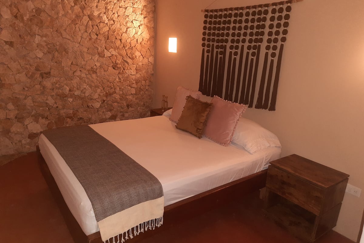 A bedroom with a stone wall, a bed with a white mattress and two pillows, a wooden nightstand, and a wall-mounted light fixture.