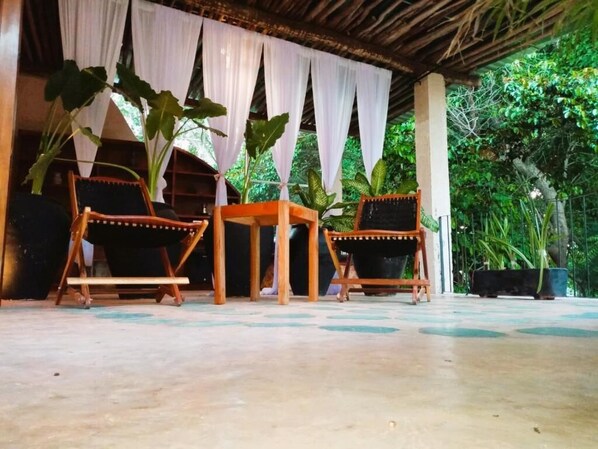 Un espacio de estar al aire libre cubierto, con dos sillas de madera y una mesa, rodeado de grandes macetas con plantas y cortinas blancas colgantes.