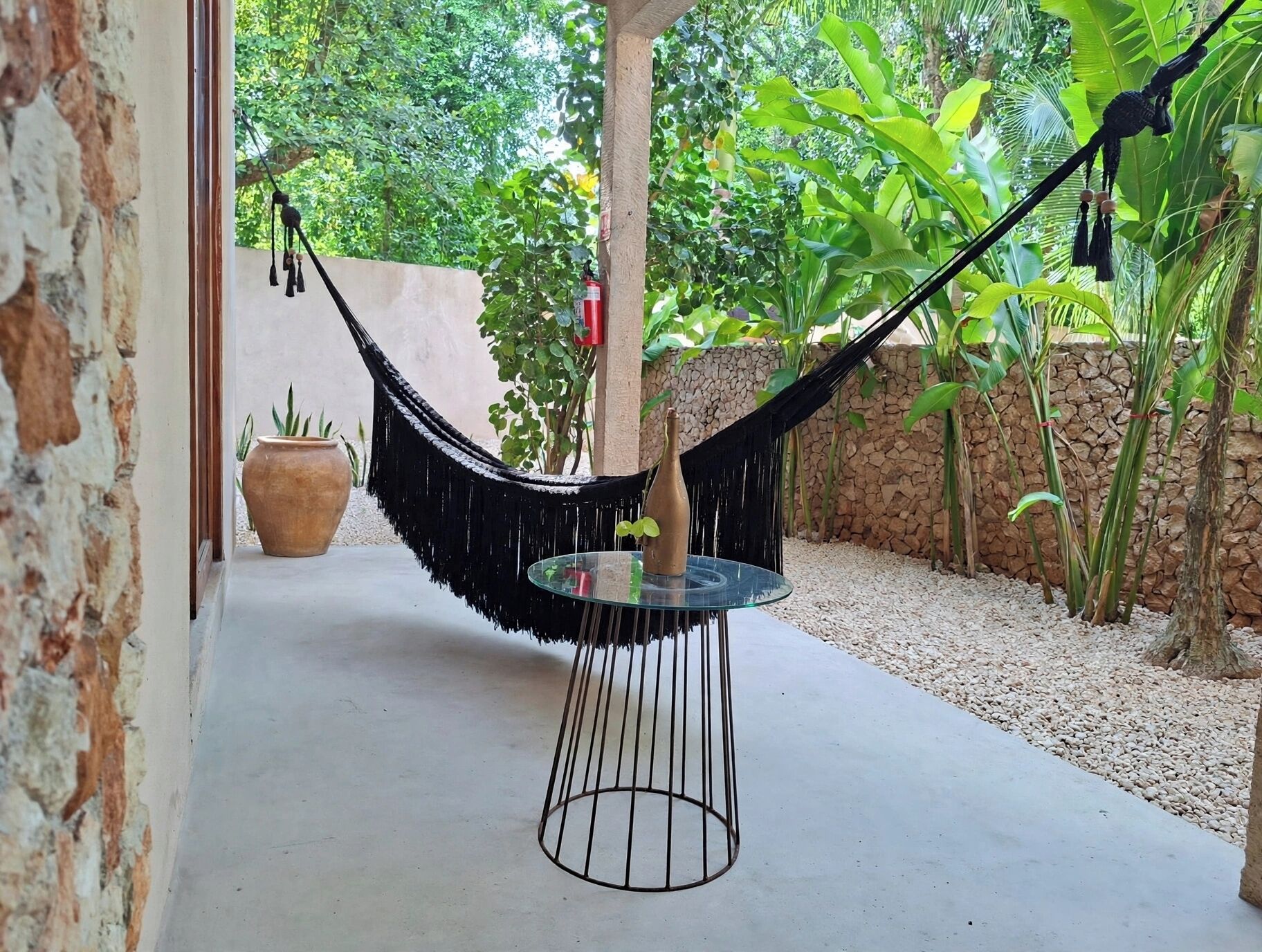 A hammock chair with a glass table, a potted plant, and a stone wall in a garden setting.