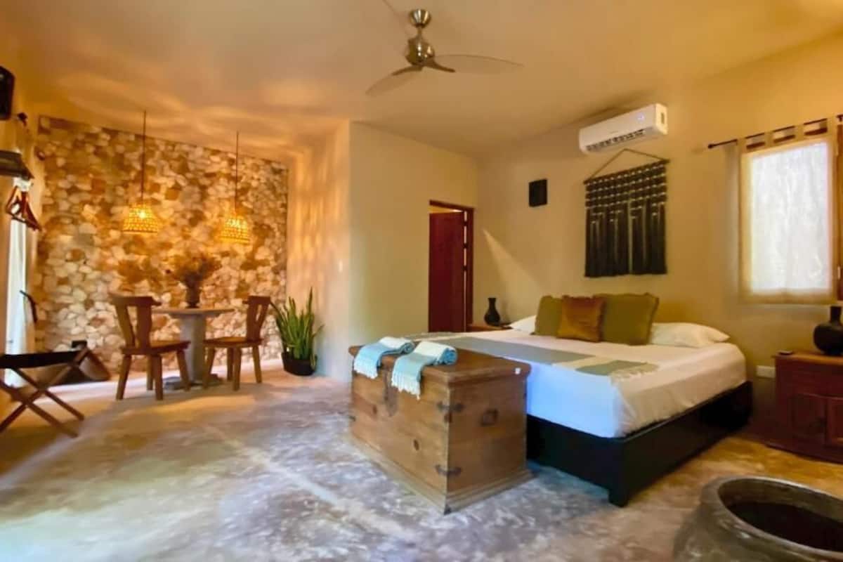A bedroom with a bed, a wooden chest, a small table with chairs, a stone wall, and a ceiling fan.