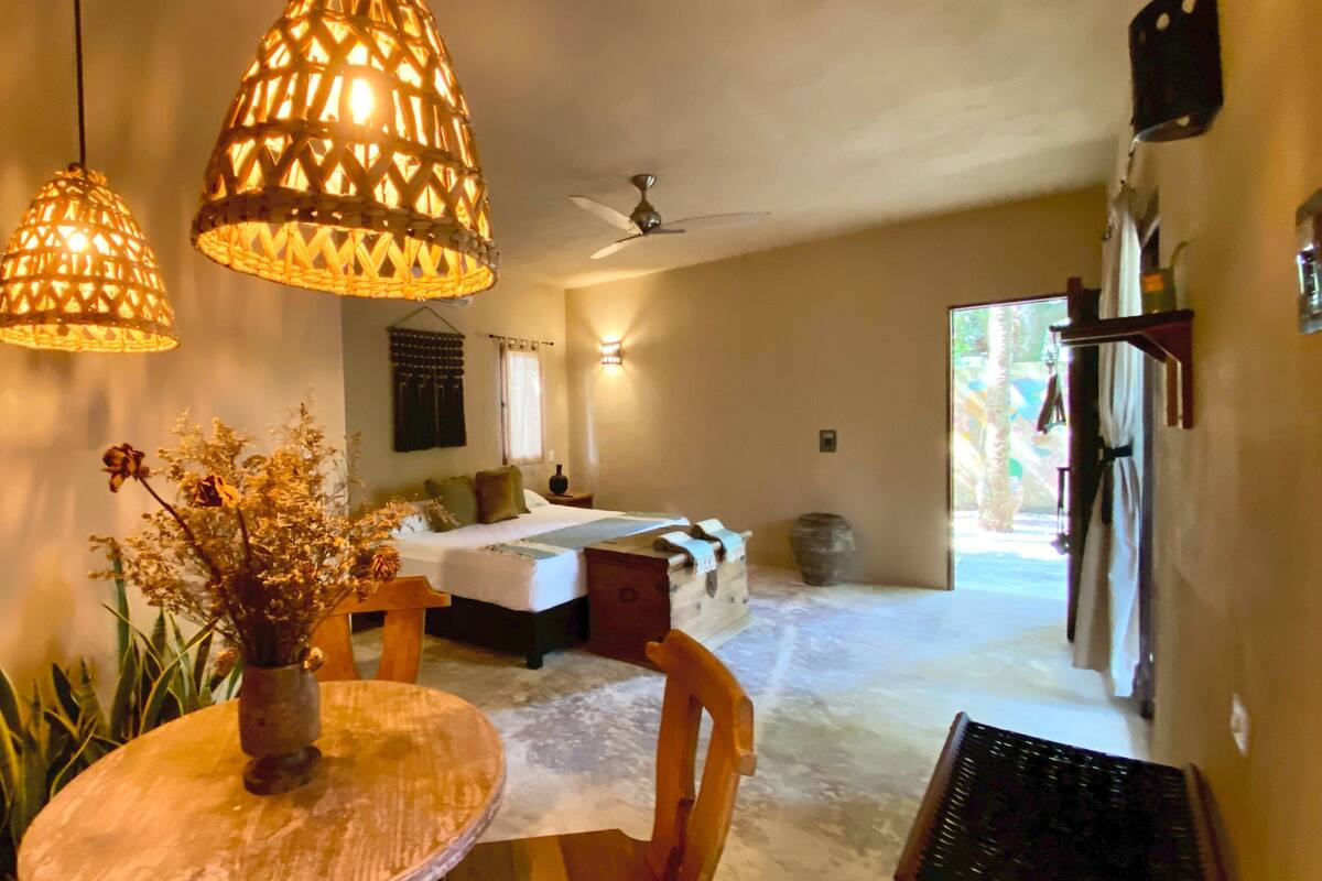 A room with a bed, a dining table with a vase of dried flowers, and hanging wicker lamps.