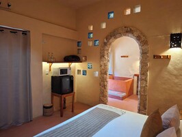Habitación de hotel con cama, escritorio con computadora, microondas y un arco de piedra que da acceso a un baño.