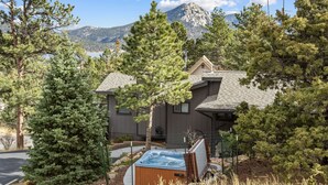 A two-story house with a hot tub in the backyard, surrounded by pine trees and a mountain in the background.