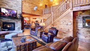 A log cabin interior with a stone fireplace, leather furniture, a staircase, and a television in the living area. - KNOTTYWOOD - Upscale, Elegant, 8X Award-Winning! - Located in pvt. gated resort. (Gatlinburg)