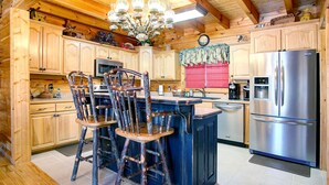 A rustic kitchen with wooden cabinets, a central island with stools, a stainless steel refrigerator, and a chandelier. - KNOTTYWOOD - Upscale, Elegant, 8X Award-Winning! - Located in pvt. gated resort. (Gatlinburg)
