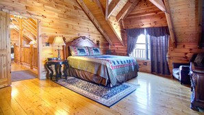 A log cabin bedroom with a wooden ceiling, a bed with a patterned bedspread, a nightstand, a lamp, and a window with curtains. - KNOTTYWOOD - Upscale, Elegant, 8X Award-Winning! - Located in pvt. gated resort. (Gatlinburg)