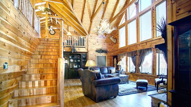A log cabin interior with a staircase, wooden walls, a fireplace, a television, and a large window. - KNOTTYWOOD - Upscale, Elegant, 8X Award-Winning! - Located in pvt. gated resort. (Gatlinburg)