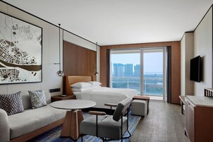 A modern hotel room with a large bed, a sofa, a dining table, and a view of the cityscape through the window.