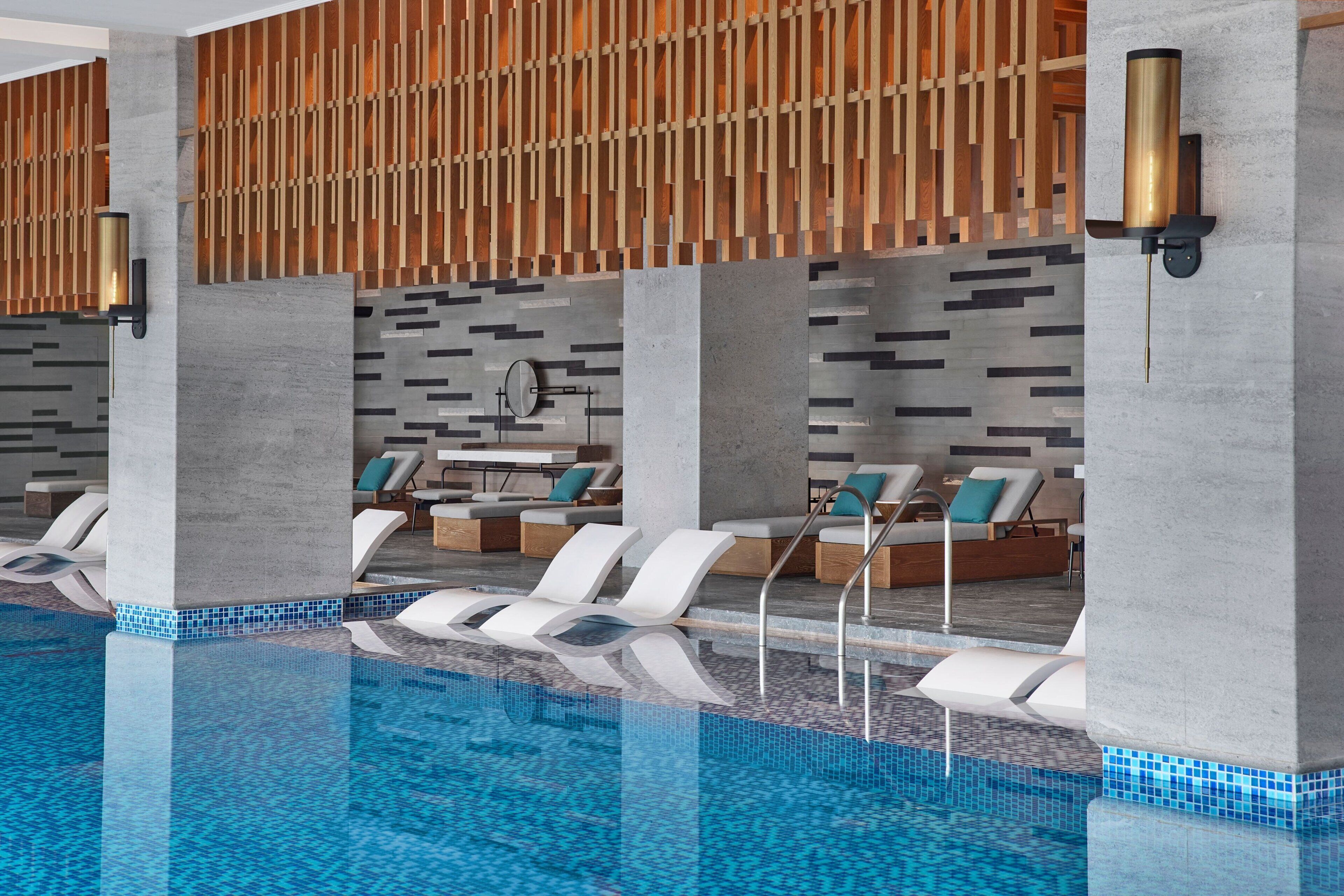 En moderne swimmingpool indendørs med liggestole og en væg af trælameller.