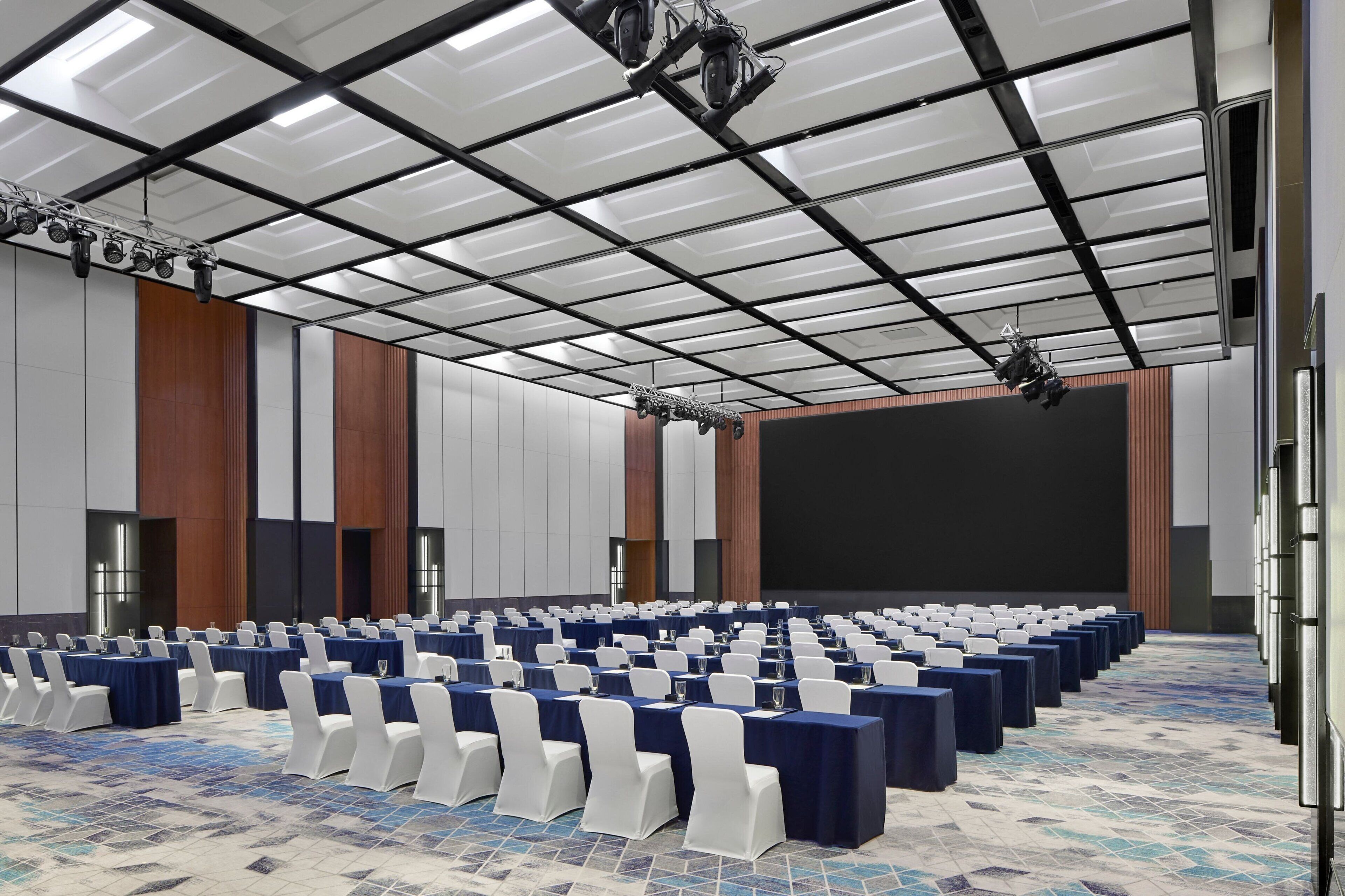 Una sala de conferencias con filas de mesas y sillas, una pantalla grande y un techo con luminarias.