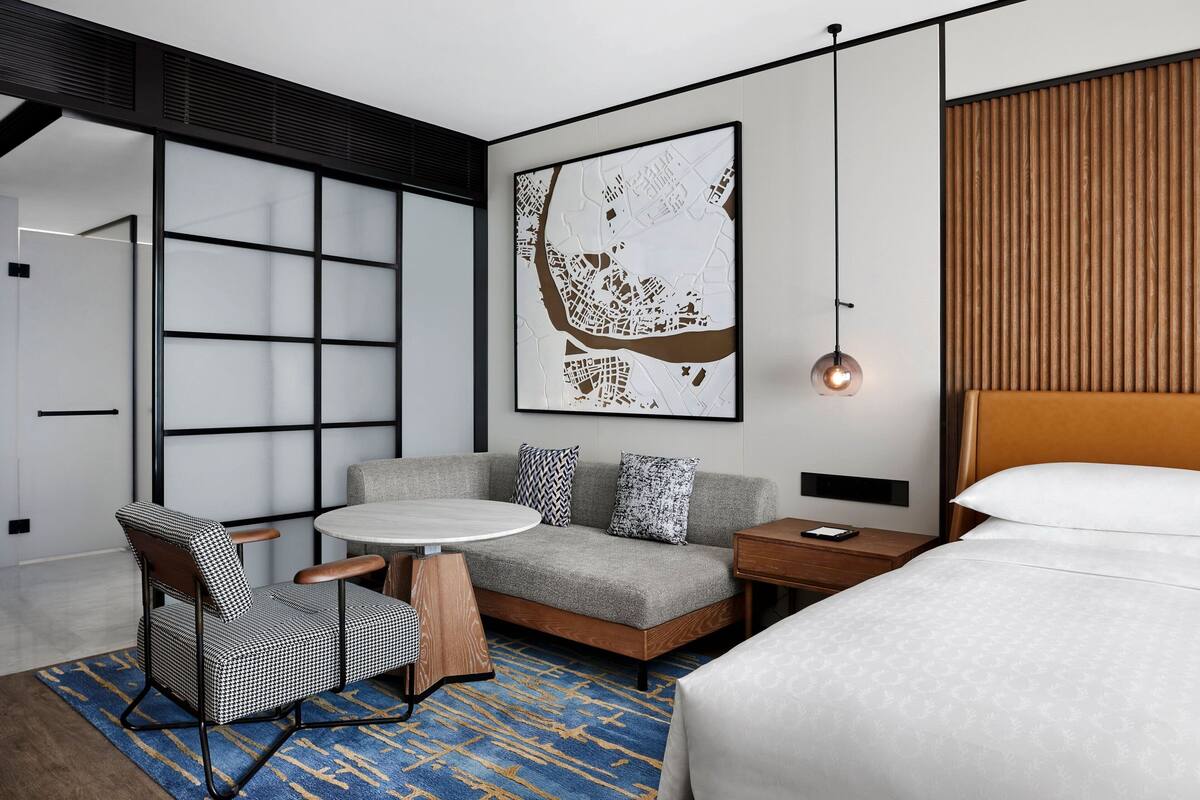 Moderni hotellihuone, jossa on sohva, pieni pöytä, tuoli, sänky ja suuri seinäkartta.