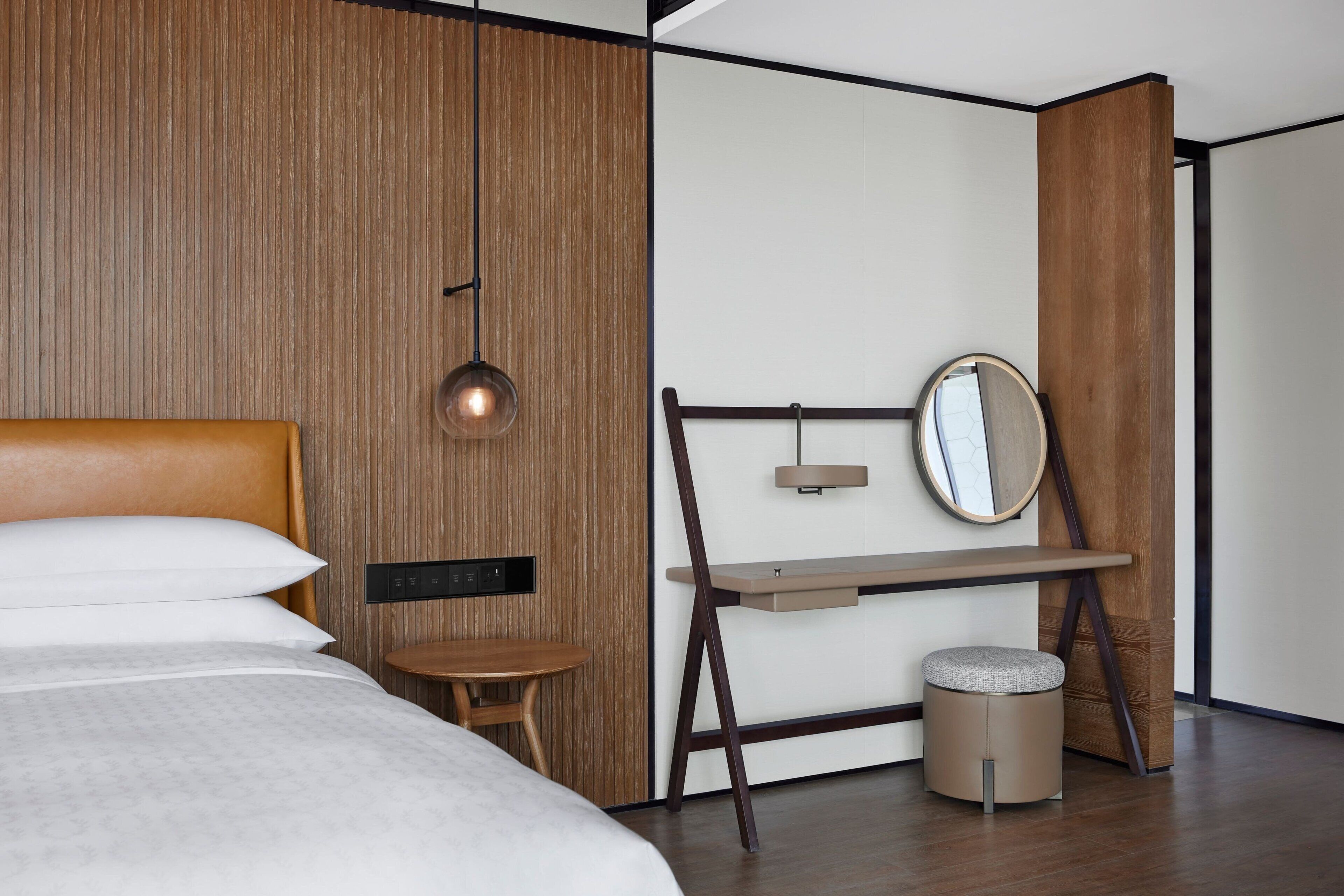 A modern hotel room with a wooden accent wall, a bed with white linens, a desk with a mirror, and a small stool.