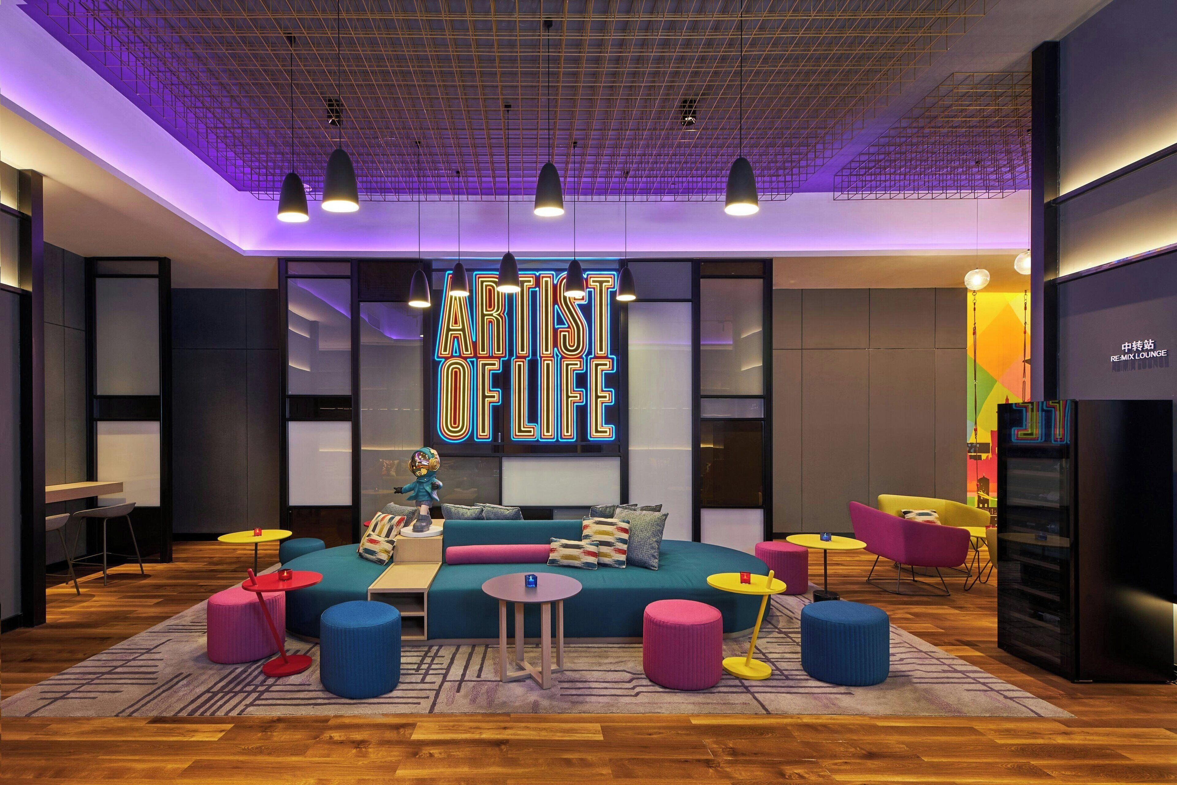 Un espacio lounge moderno con un letrero de neón que dice "ARTIST OF LIFE", que incluye un sofá, otomanas coloridas e iluminación colgante.