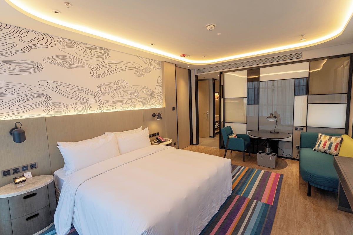 Une chambre d’hôtel moderne avec un grand lit, un bureau et une chaise. On y trouve une petite table avec une lampe, un miroir et un tapis coloré.