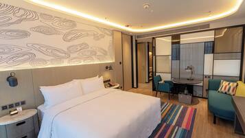 Quarto de hotel moderno com uma cama grande, uma escrivaninha e uma cadeira. Há uma pequena mesa com uma luminária, um espelho e um tapete colorido.