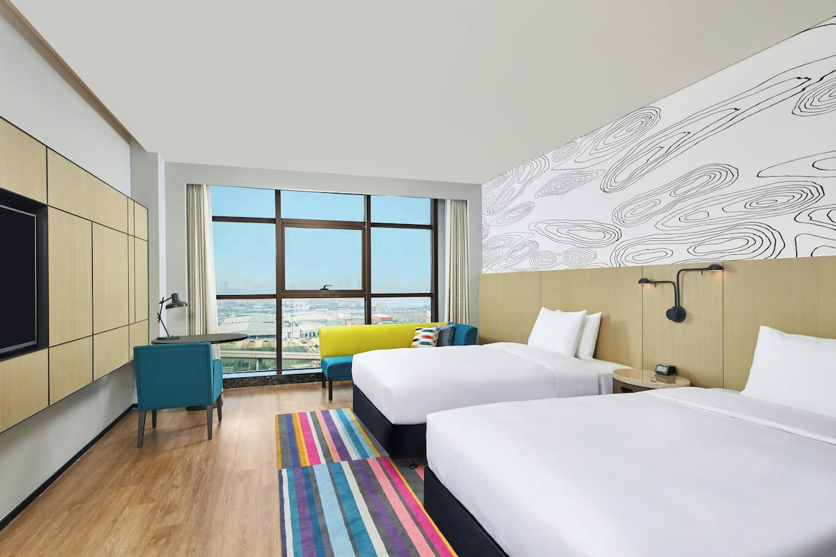 Une chambre d’hôtel avec deux lits, un bureau, une chaise, un tapis rayé aux couleurs vives et une grande fenêtre donnant sur la ville.