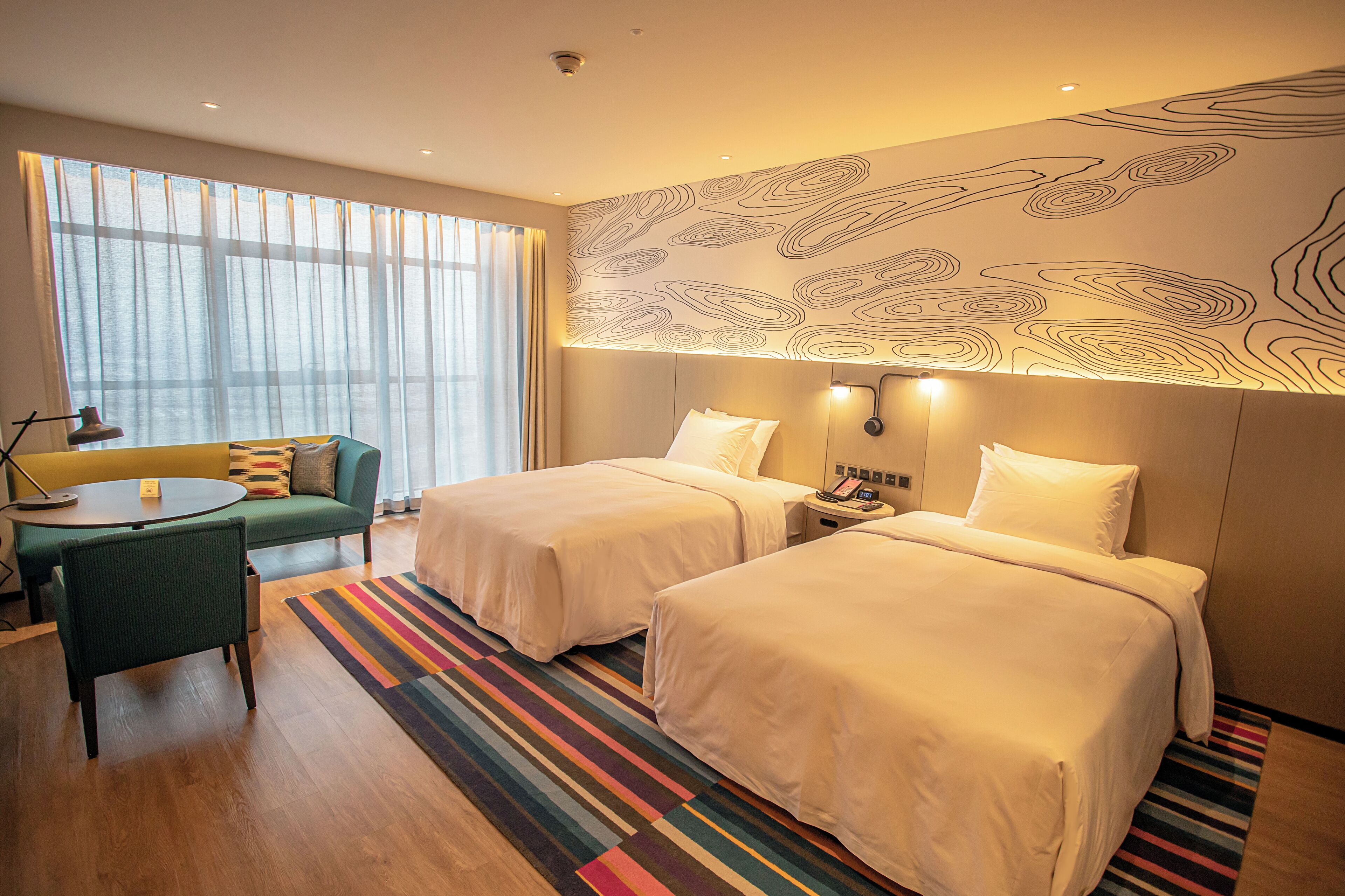 Een hotelkamer met twee bedden, een bank, een klein tafeltje en een groot raam met gordijnen.