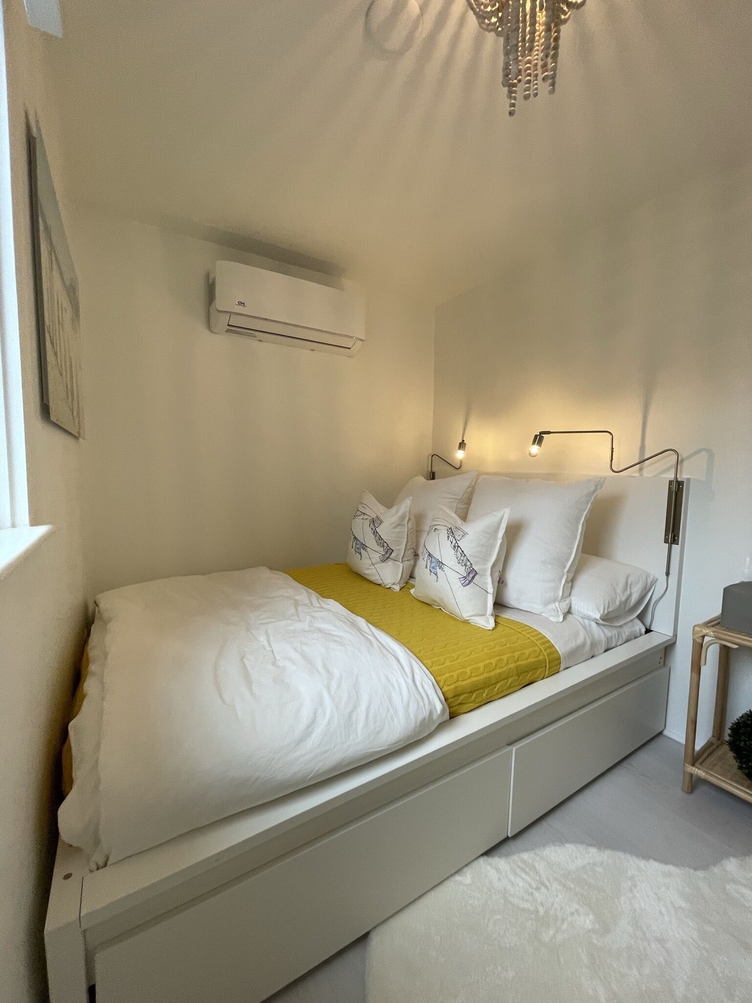 A bed with white bedding, a yellow throw, and decorative pillows. There is a bedside table with a lamp, and an air conditioning unit on the wall.
