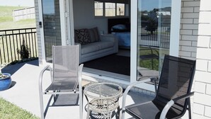 An outdoor patio with a sliding glass door leading to a living area. There are two chairs and a small round table. - Paradise on the Sand (Whakatane)