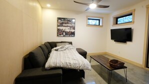 A modern living room with a sofa, a flat-screen TV, a ceiling fan, and a window with a view of trees. - Private Suite & Hot Tub @ Red (Rossland)