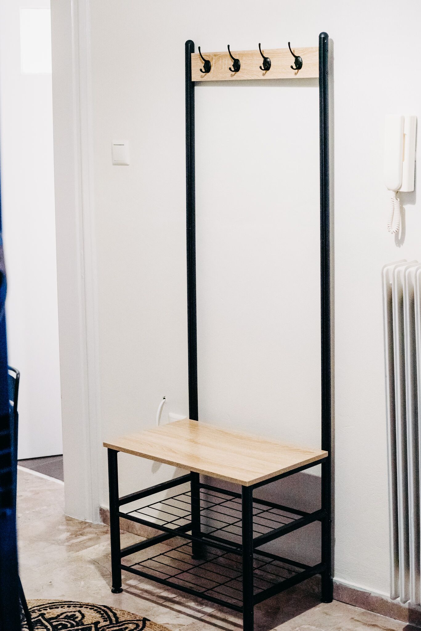 Une étagère haute et étroite, en métal noir, avec une tablette supérieure en bois clair, le tout adossé à un mur blanc.
