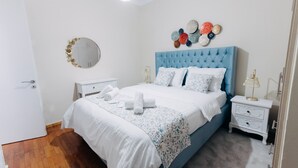 Un dormitorio ordenado con una cabecera tapizada en azul, ropa de cama blanca, un espejo redondo y una mesita de noche con una lámpara.