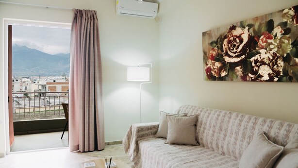 Una sala de estar con un sofá, un cuadro de flores, una ventana con vistas a edificios y montañas, y una cortina.