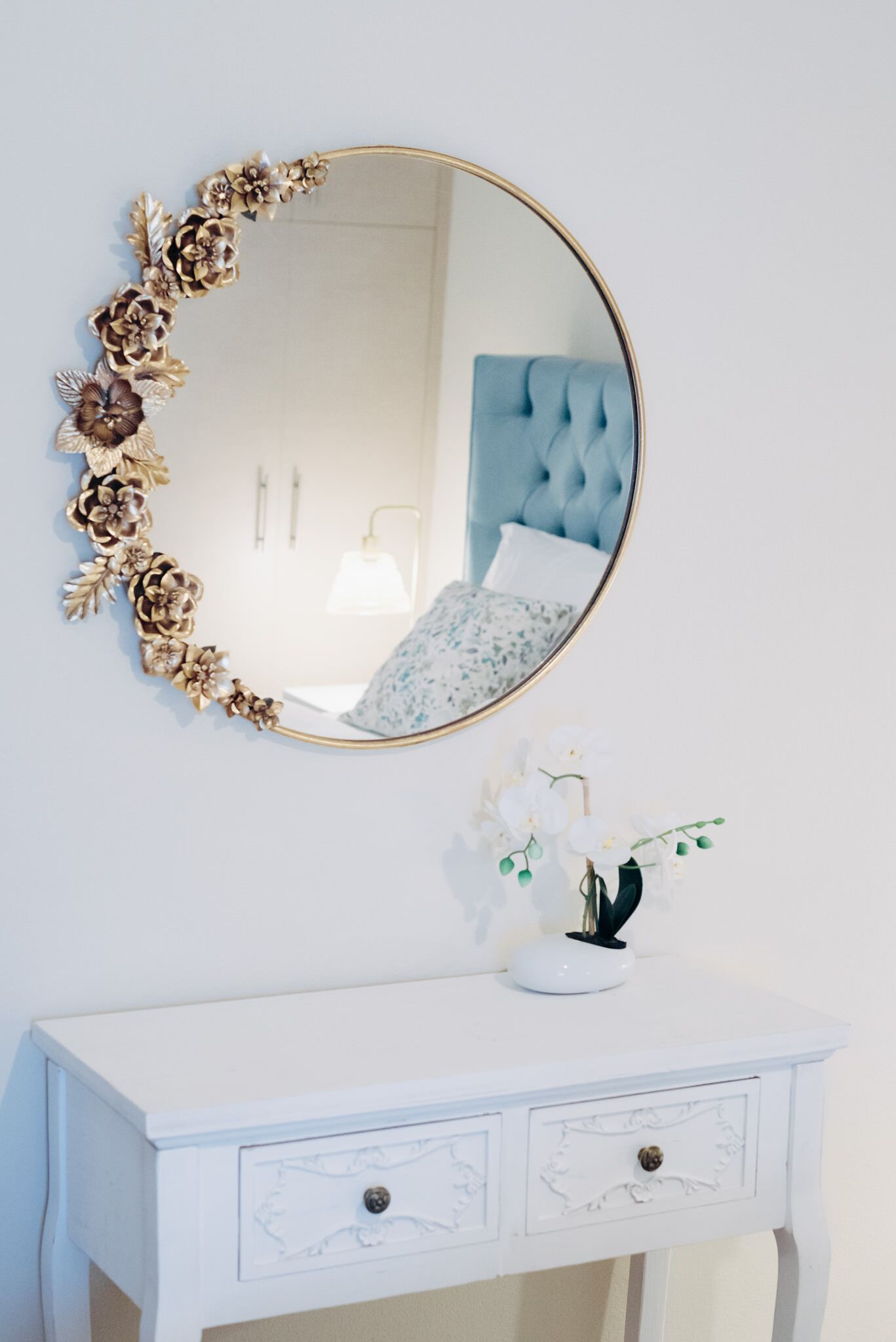 A white dresser with a gold-framed mirror and a small white vase with flowers.