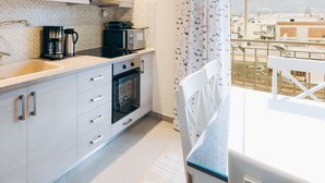 Cocina moderna con gabinetes blancos, cafetera, microondas y horno. Hay un comedor con mesa y sillas, y se ve edificios y montañas a través del ventanal.