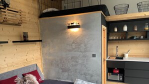 A modern bedroom with a bed, a bedside table, a wall-mounted shelf with decorative items, and a small kitchenette area. - View and Location! (Morzine)