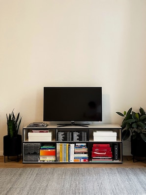 Un téléviseur à écran plat, posé sur un meuble noir, avec une bibliothèque en dessous contenant divers livres et DVD.