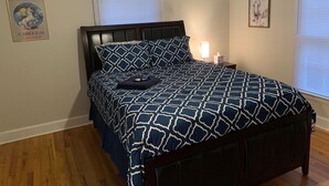 Une chambre à coucher avec un lit en bois foncé, une couvre-lit à motifs, une table de chevet avec une lampe et deux fenêtres équipées de stores.