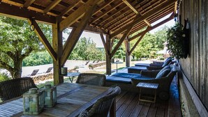 Un espacio de estar al aire libre techado, con mesas de madera y sillas de mimbre, con vistas a una piscina y follaje.