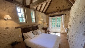 Un dormitorio con una cama grande, un escritorio de madera y una silla. Hay dos ventanas con cortinas, una puerta con paneles de vidrio y una pared de piedra.
