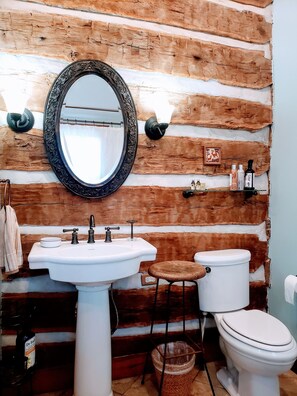 A rustic bathroom with a wooden wall, a round mirror, a white sink, a toilet, and a stool.
